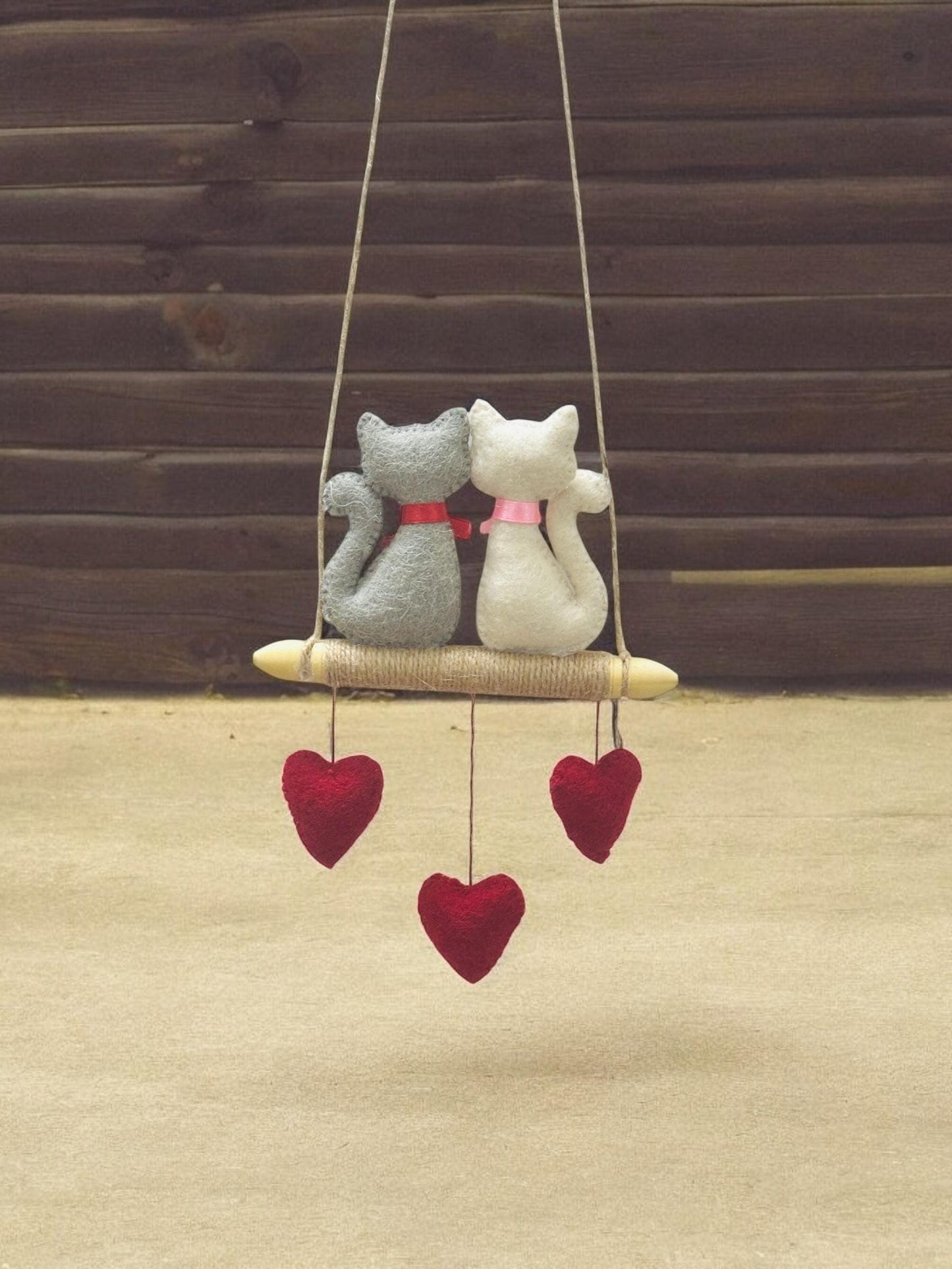 Decorative hanging swing with two cat-shaped plush toys and heart decorations on a wooden background.