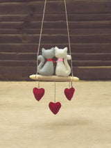 Decorative hanging swing with two cat-shaped plush toys and heart decorations on a wooden background.