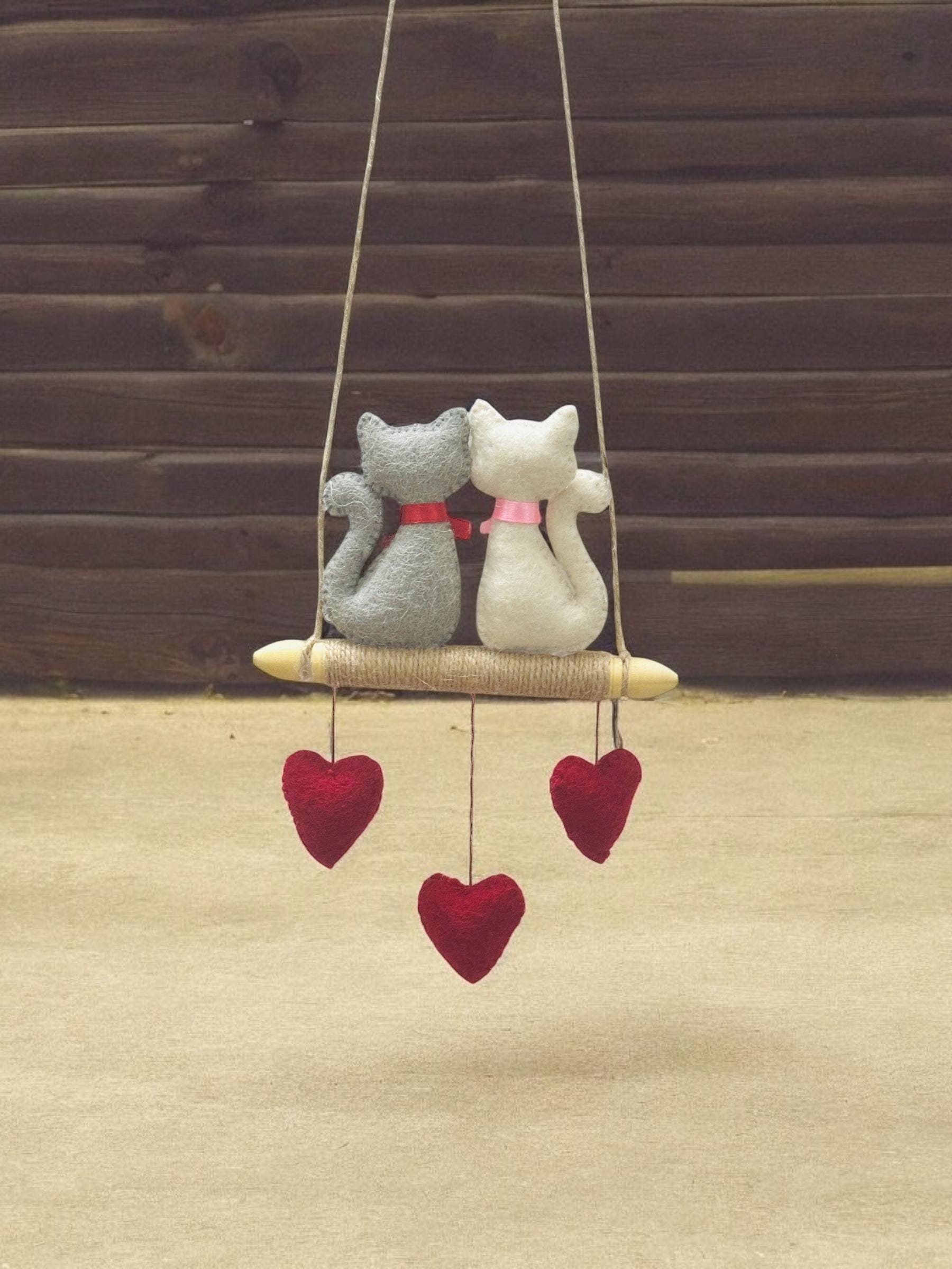 Decorative hanging swing with two cat-shaped plush toys and heart decorations on a wooden background.