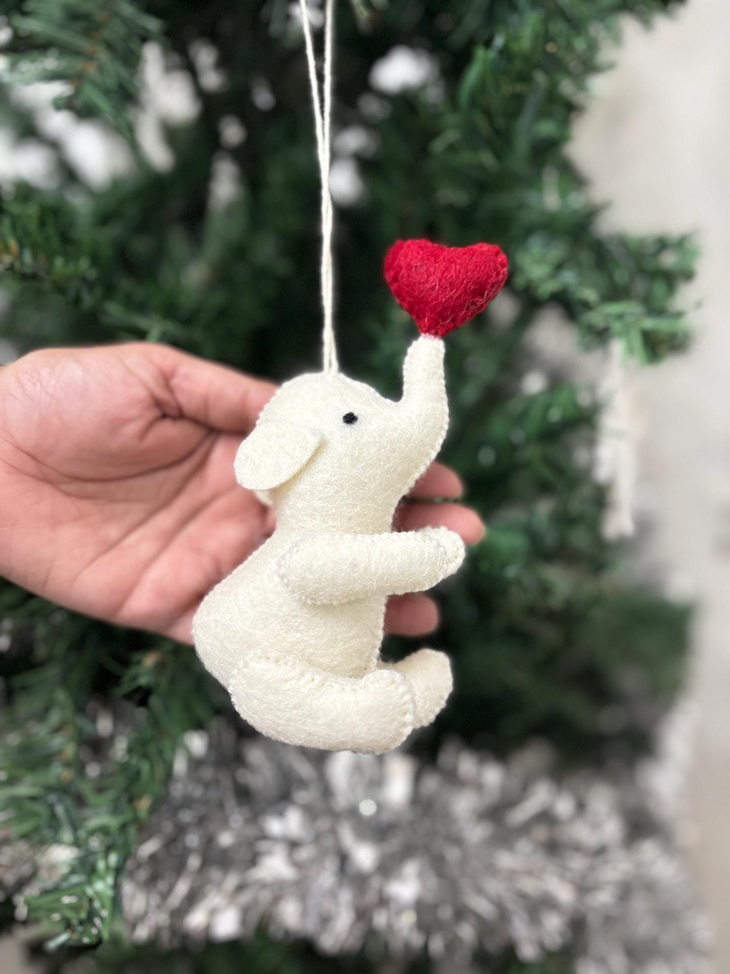 White elephant ornament with a red heart held in front of a decorated Christmas tree.