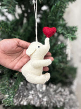 White elephant ornament with a red heart held in front of a decorated Christmas tree.