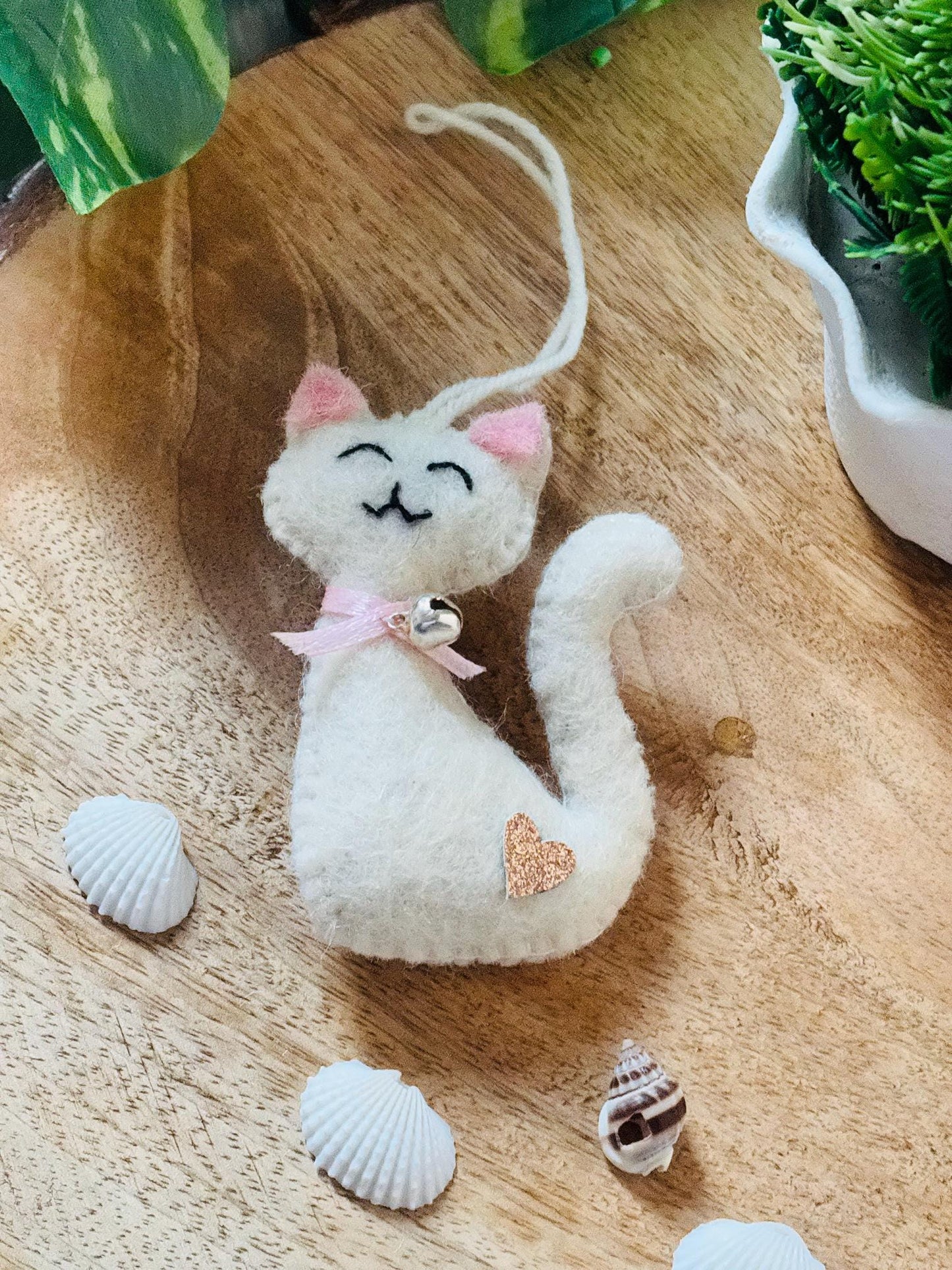White cat-shaped plush toy with pink bow on a wooden surface with shells and plants.