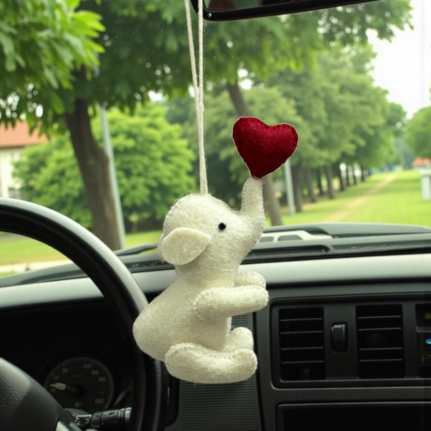 White plush elephant with a red heart hanging from a car's rearview mirror with a green outdoor background.