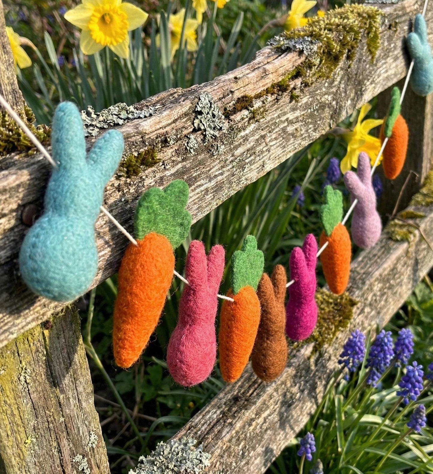Decorative string of felted Easter bunnies and carrots hanging on a wooden fence with flowers in the background.