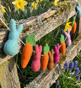 Decorative string of felted Easter bunnies and carrots hanging on a wooden fence with flowers in the background.