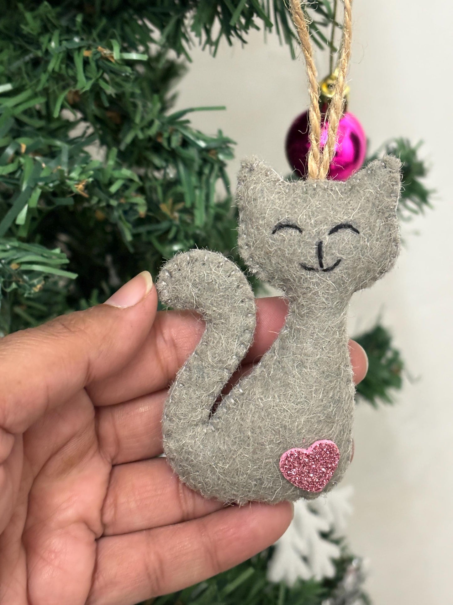 Gray cat-shaped ornament with a pink heart on a hand, Christmas tree in the background