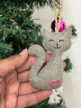 Gray cat-shaped ornament with a pink heart on a hand, Christmas tree in the background