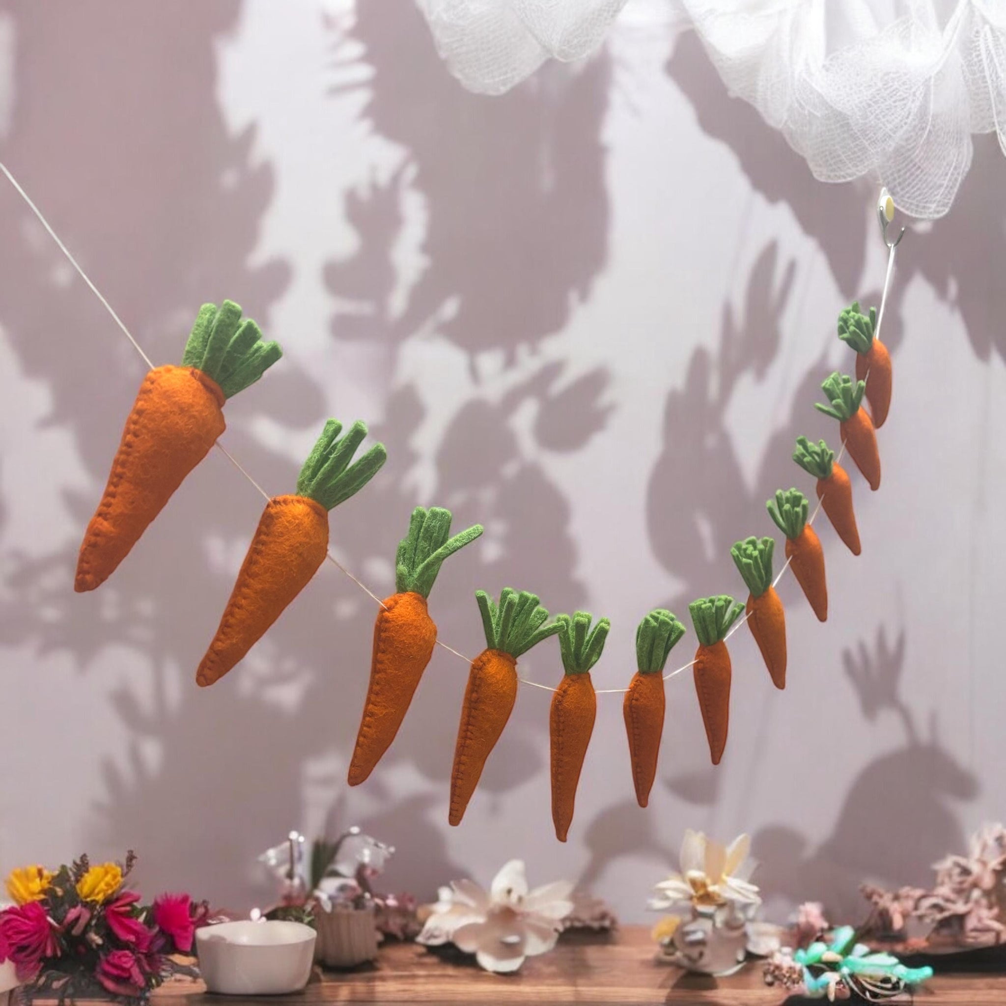 Felt carrot garland hanging against a light background with decorative items on a table below.
