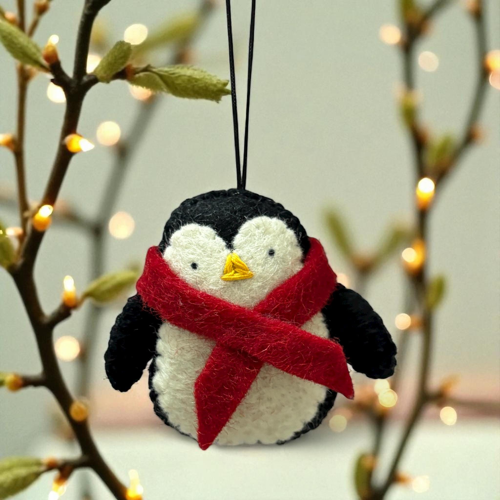 Felt penguin ornament with a red scarf hanging on a branch with blurred lights in the background