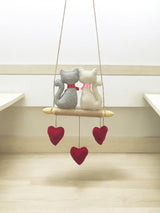 Decorative hanging with two cat-shaped figures and red heart shapes on a light wooden floor.