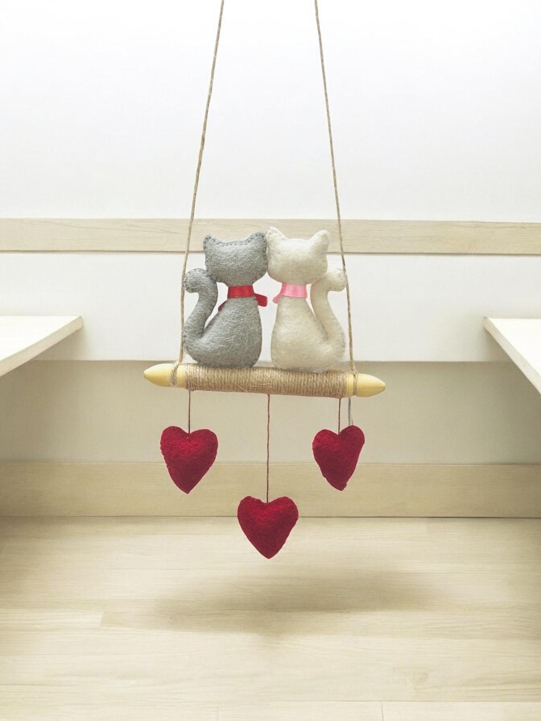 Decorative hanging with two cat-shaped figures and red heart shapes on a light wooden floor.