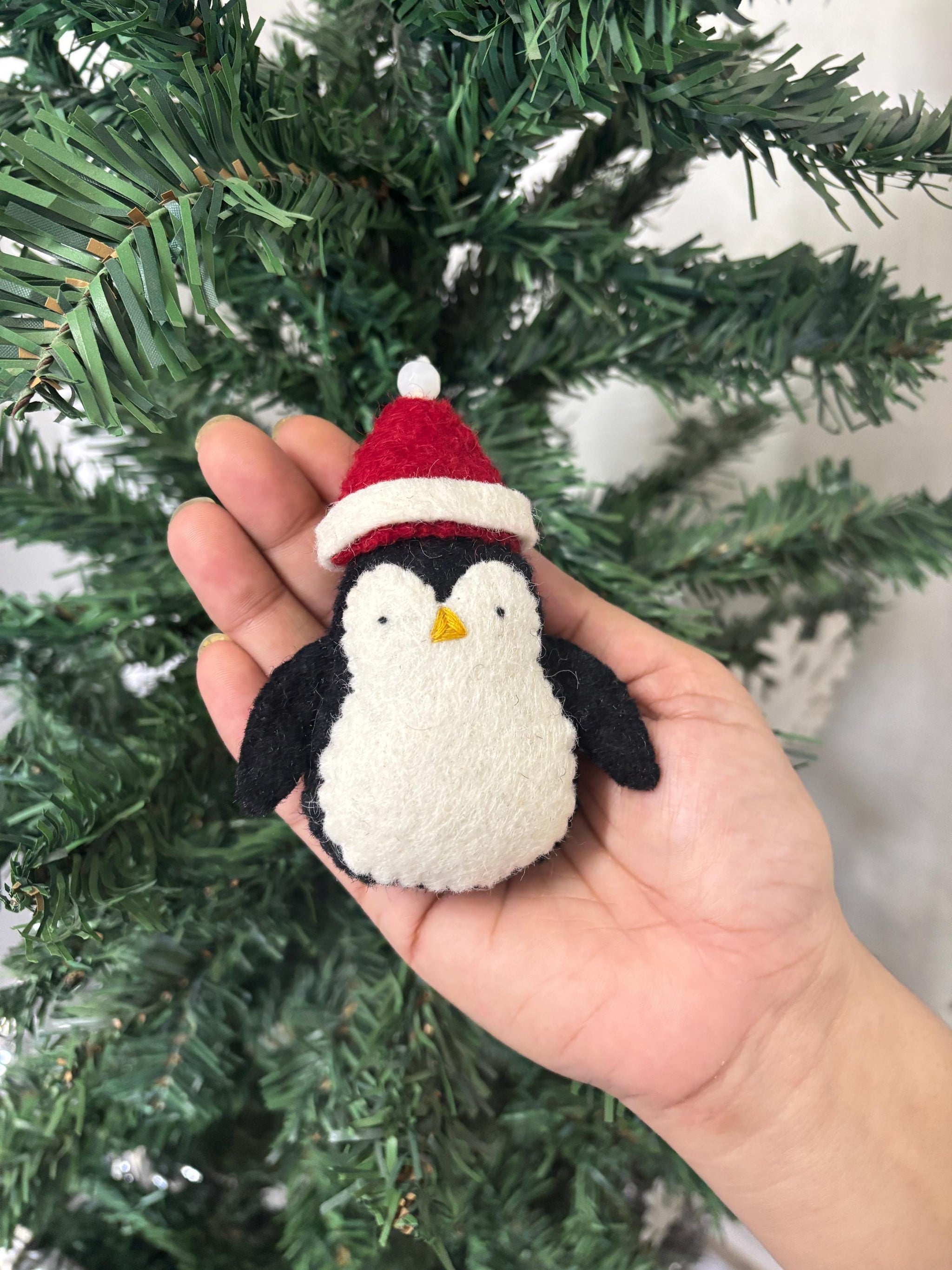 Hand holding a small crocheted penguin ornament with a Santa hat in front of a Christmas tree.