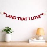 Decorative banner with 'LAND THAT I LOVE' on a wall above a wooden shelf with a plant, books, and lamp.