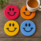 Four smiley face coasters in red, orange, yellow, and blue on a wooden surface with a cup of tea.