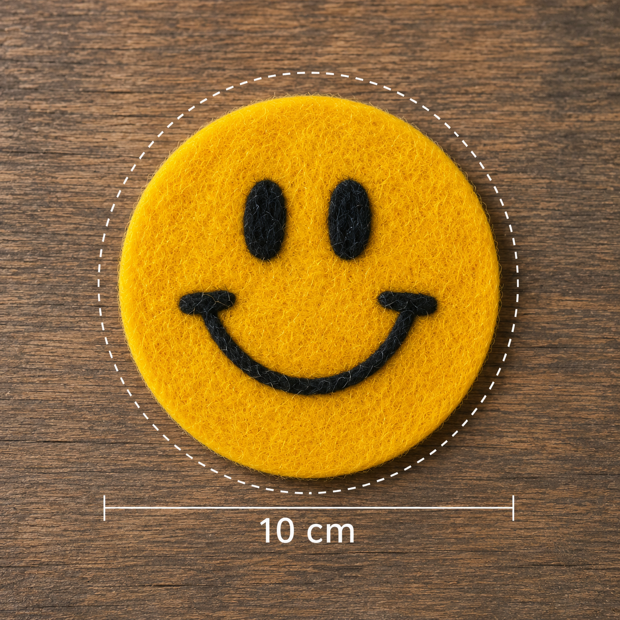 Yellow smiley face patch with black details on a wooden surface, showing measurement of 10 cm.