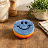 Multicolored smiley face scrubber on a wooden surface with a plant and jars in the background