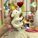 Hand holding a small white elephant plush toy with a red heart on a blurred background of toys.