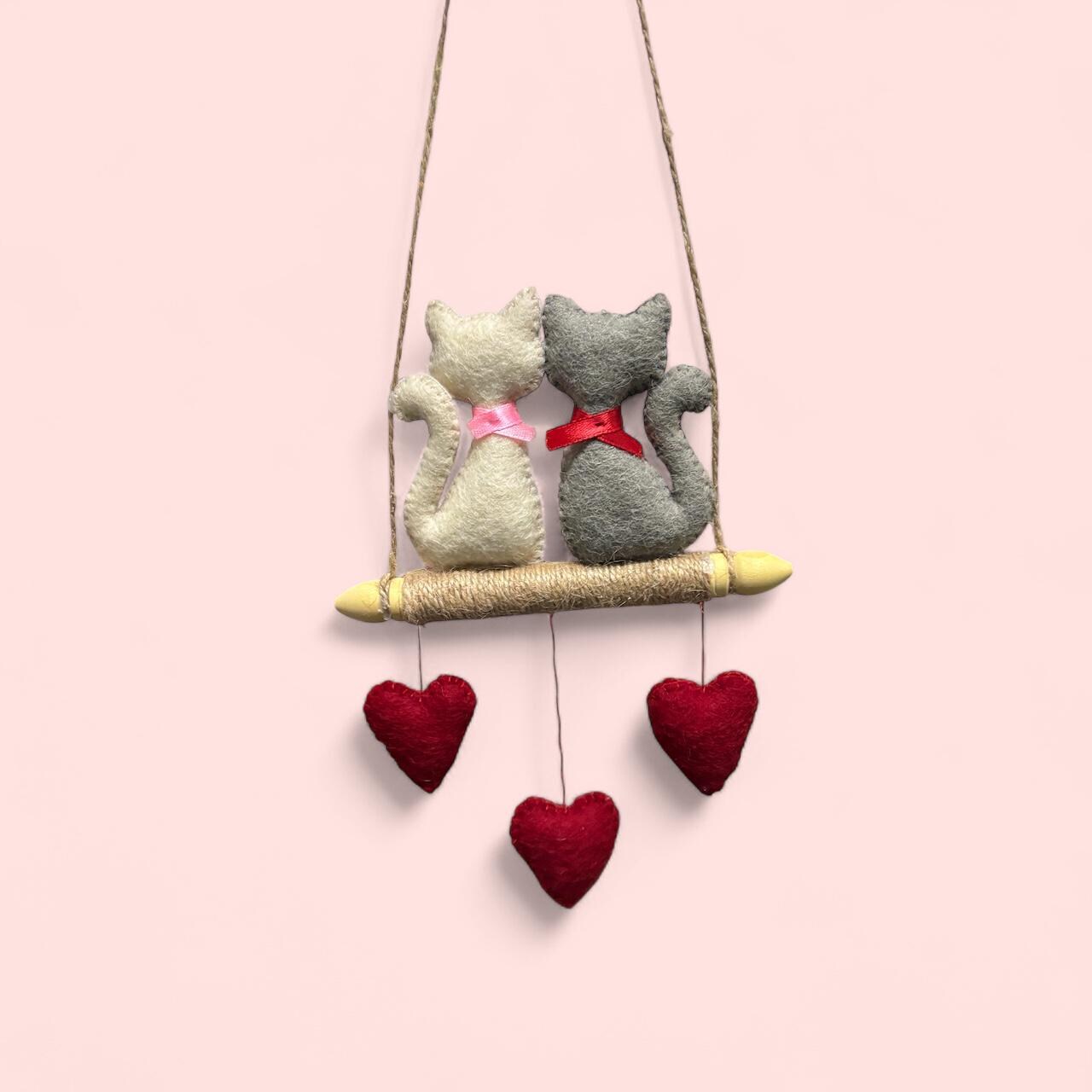 Decorative hanging with two cats on a swing and three hearts below against a pink background