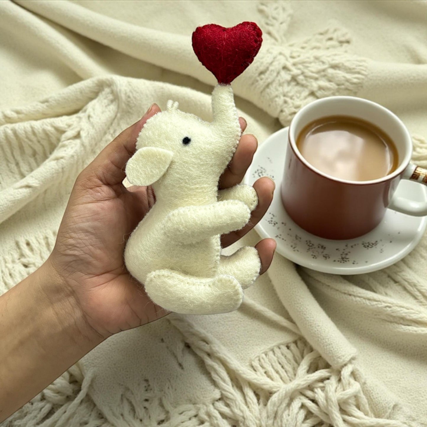 Hand holding a small plush elephant with a red heart above it, next to a cup of coffee on a white blanket.