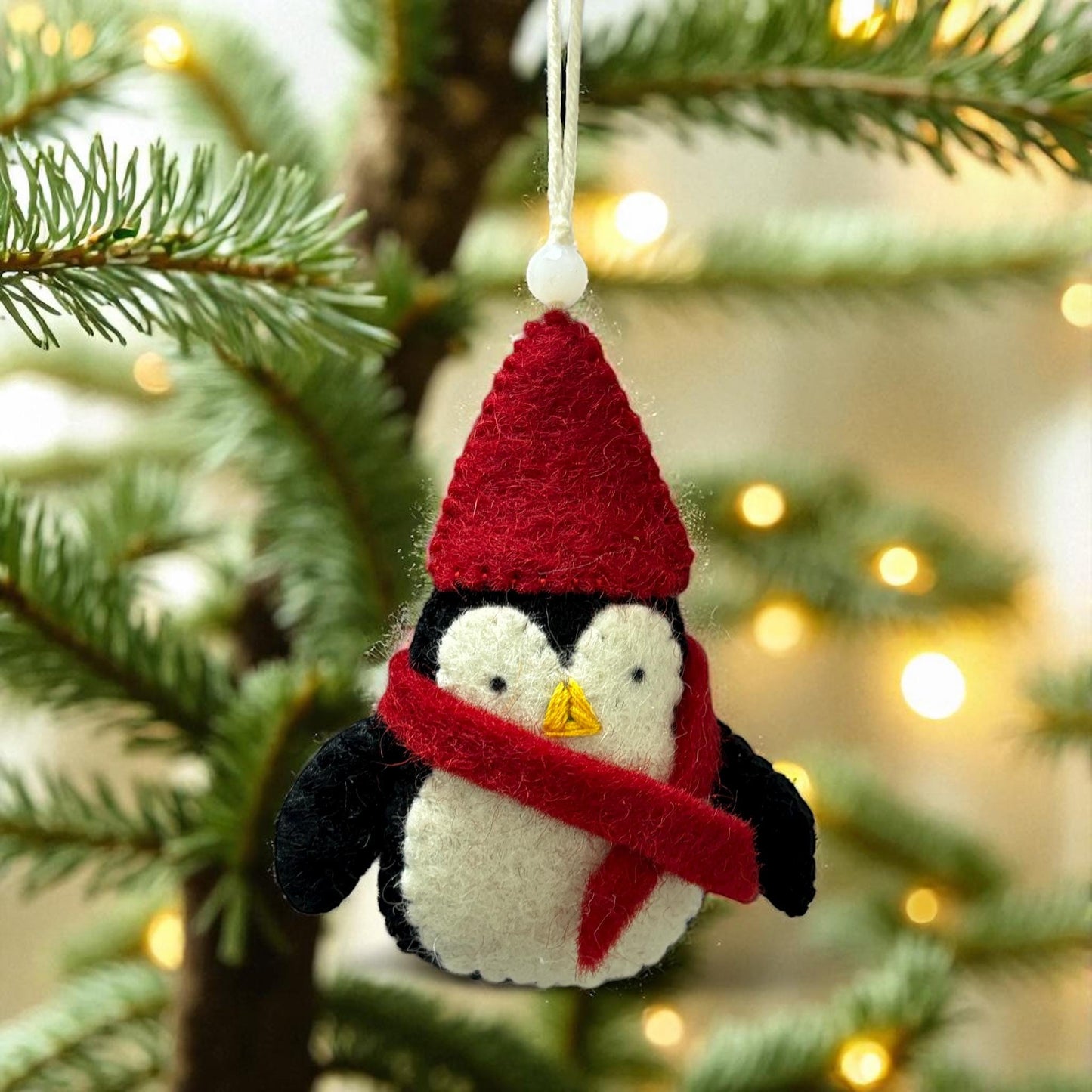 Felt penguin ornament with a red hat and scarf hanging on a Christmas tree.