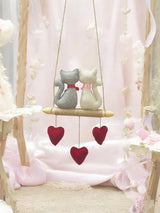 Decorative swing with cat figures and heart-shaped ornaments against a soft pink background